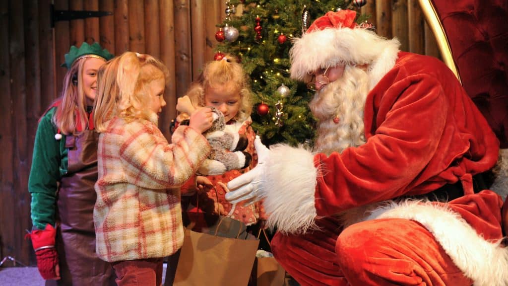 Two young girls meeting with Santa, as he hands one of them a plush reindeer toy.